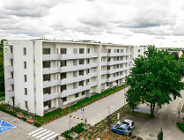 Zdjęcie przedstawia nowoczesny, czteropiętrowy budynek mieszkalny o białej elewacji. Charakterystycznym elementem są długie balkony z jasnymi, ażurowymi osłonami. Przed blokiem znajduje się czysty asfaltowy podjazd, przejście dla pieszych oraz wyznaczone niebieską farbą miejsce parkingowe dla osób z niepełnosprawnościami. Wokół widać młode drzewa i starannie utrzymaną zieleń, a w tle gęsty las pod pochmurnym niebem.