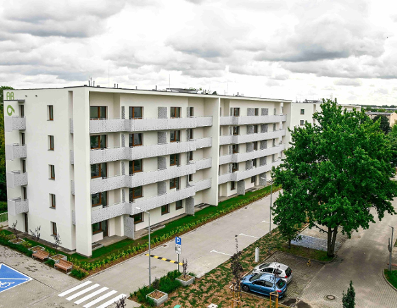 Zdjęcie przedstawia nowoczesny, czteropiętrowy budynek mieszkalny o białej elewacji. Charakterystycznym elementem są długie balkony z jasnymi, ażurowymi osłonami. Przed blokiem znajduje się czysty asfaltowy podjazd, przejście dla pieszych oraz wyznaczone niebieską farbą miejsce parkingowe dla osób z niepełnosprawnościami. Wokół widać młode drzewa i starannie utrzymaną zieleń, a w tle gęsty las pod pochmurnym niebem.