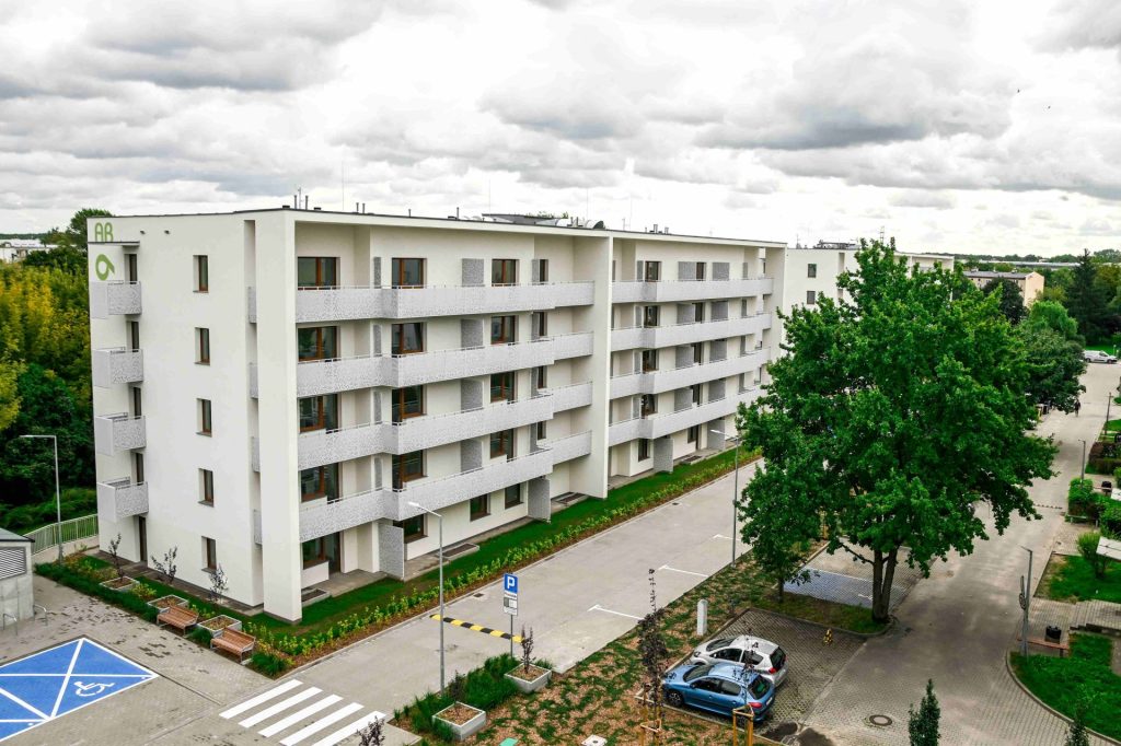 Zdjęcie przedstawia nowoczesny, pięciokondygnacyjny budynek mieszkalny o białej elewacji, wyposażony w liczne balkony z jasnymi, ażurowymi balustradami oraz brązową stolarkę okienną. Przed blokiem znajduje się uporządkowana infrastruktura: asfaltowa droga wewnętrzna, miejsca parkingowe (w tym wyraźnie oznakowane niebieskie miejsce dla osób z niepełnosprawnościami) oraz niska zieleń i młode drzewa.