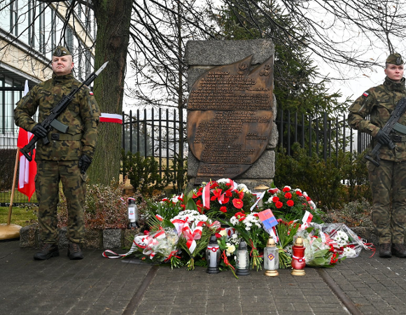 Zdjęcie przedstawia obelisk przed którym leżą kwiaty i zcznicze. Obok stoi też dwóch żołnierzy.