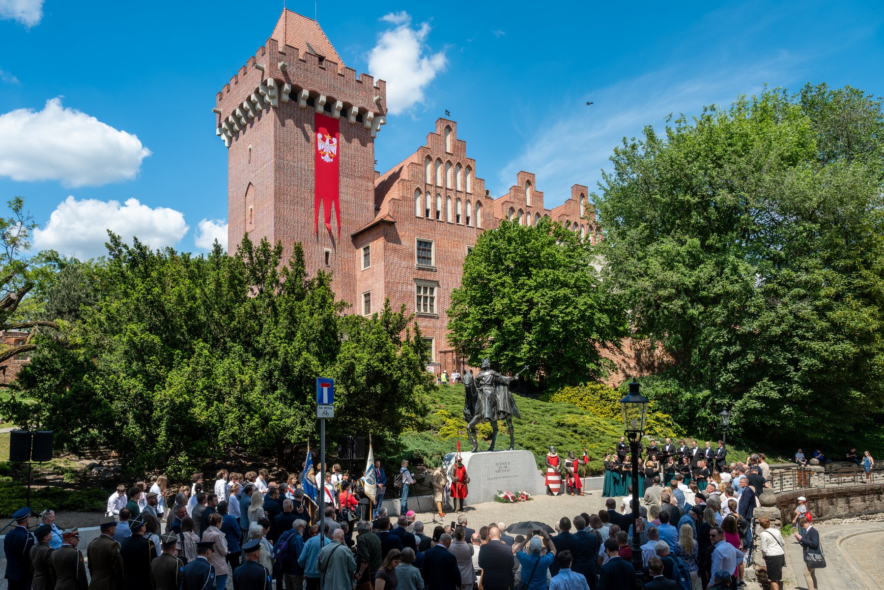 Zdjęcie przedstawia oficjalną uroczystość przed Zamkiem Królewskim w Poznaniu. Na placu przed konnym pomnikiem Przemysła II zgromadził się tłum ludzi, w tym osoby w mundurach oraz poczty sztandarowe.