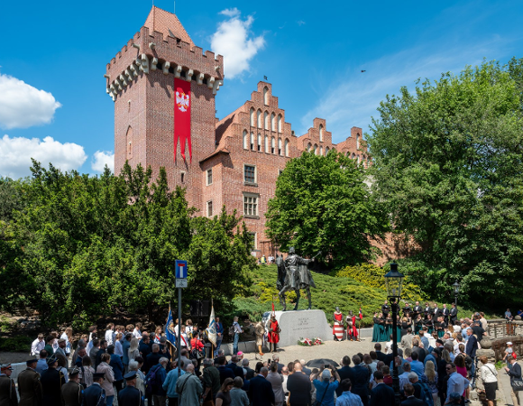 Zdjęcie przedstawia oficjalną uroczystość przed Zamkiem Królewskim w Poznaniu. Na placu przed konnym pomnikiem Przemysła II zgromadził się tłum ludzi, w tym osoby w mundurach oraz poczty sztandarowe.
