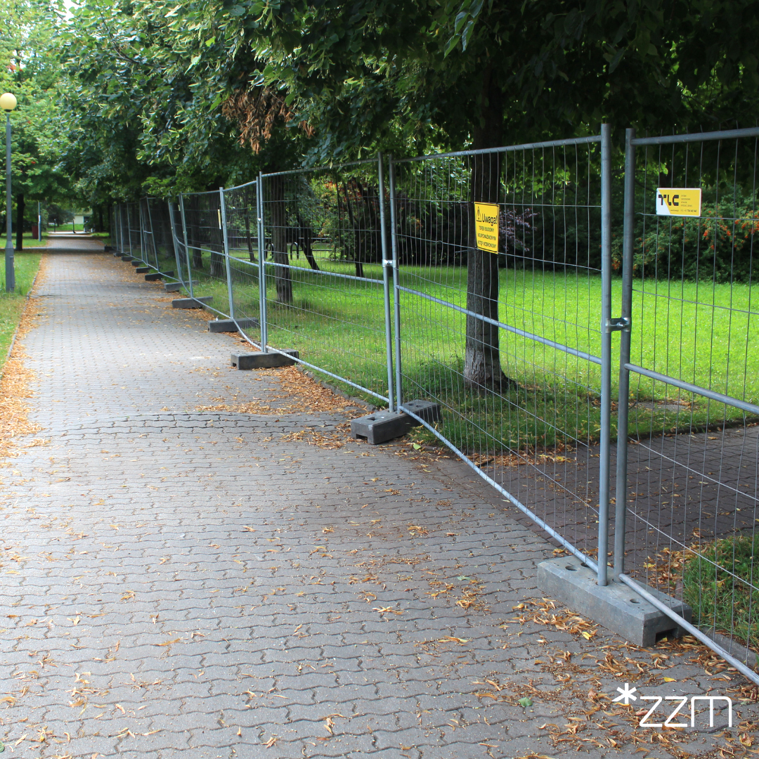 Zdjęcie przedstawia ogrodzenie, na kórym zawieszona jest tabliczka z napisem "Uwaga! Teren budowy nieupoważnionym wstęp wzbroniony". Za ogrodzeniem znajduje się park.