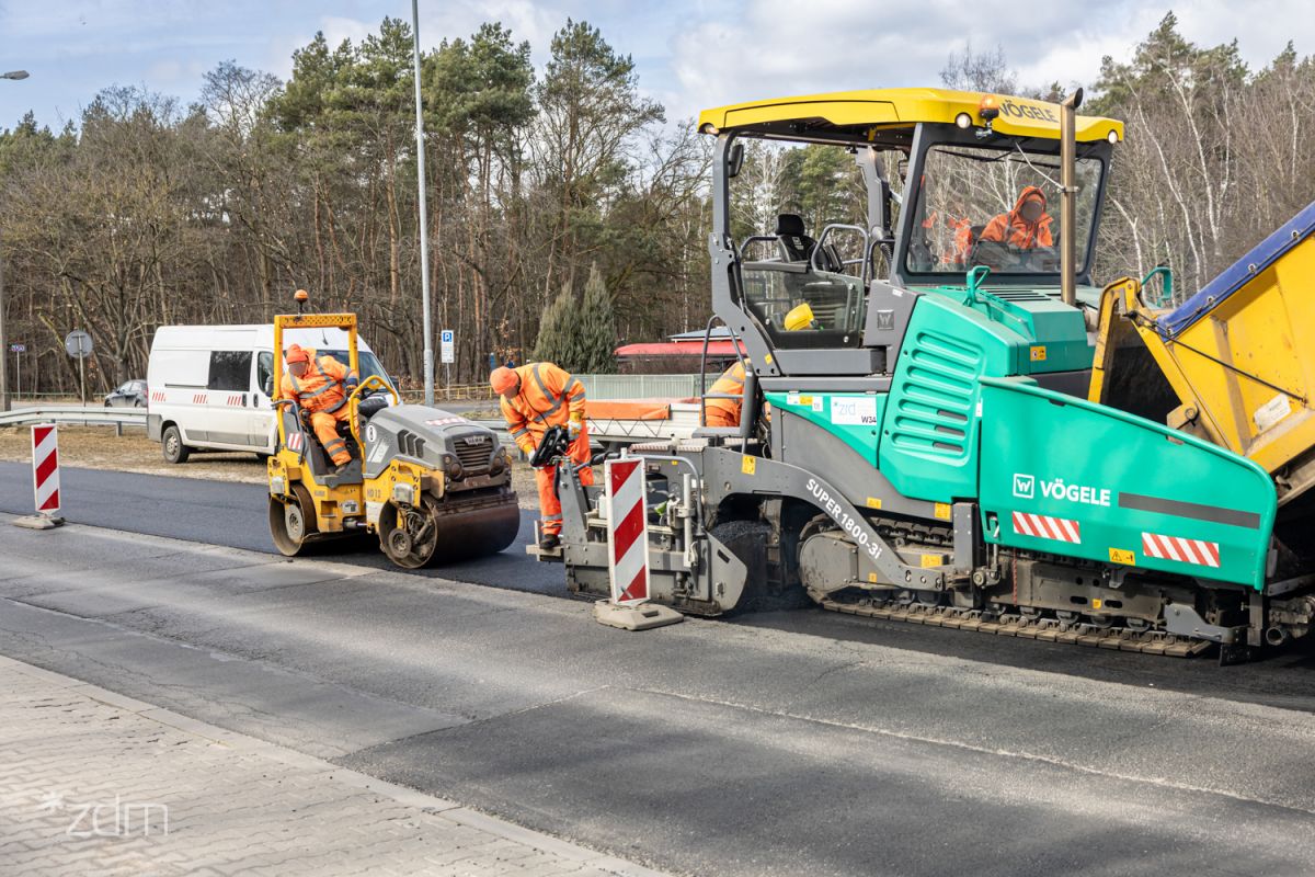 Zdjęcie przedstawia prace drogowe związane z układaniem nowej nawierzchni asfaltowej. Na pierwszym planie widać dużą, zielono-żółtą rozściełacz do asfaltu marki Vögele, obsługiwaną przez pracowników w pomarańczowych strojach ostrzegawczych.