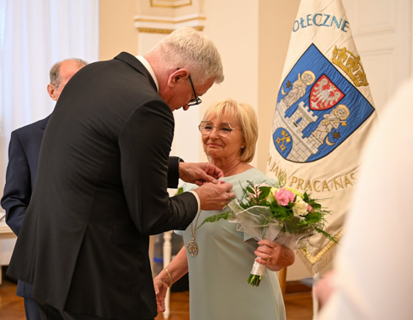 Zdjęcie przedstawia prezydenta przypinającego medal kobiecie. Obok widać flagę z herbem Poznania.