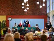Zdjęcie przedstawia prezydenta z mikrofonem stojącego za stołem oraz ludzi na krzesłach w sali gimnastycznej.