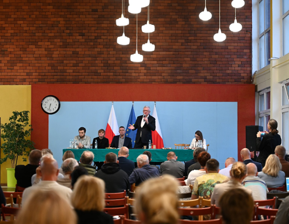 Zdjęcie przedstawia prezydenta z mikrofonem stojącego za stołem oraz ludzi na krzesłach w sali gimnastycznej.