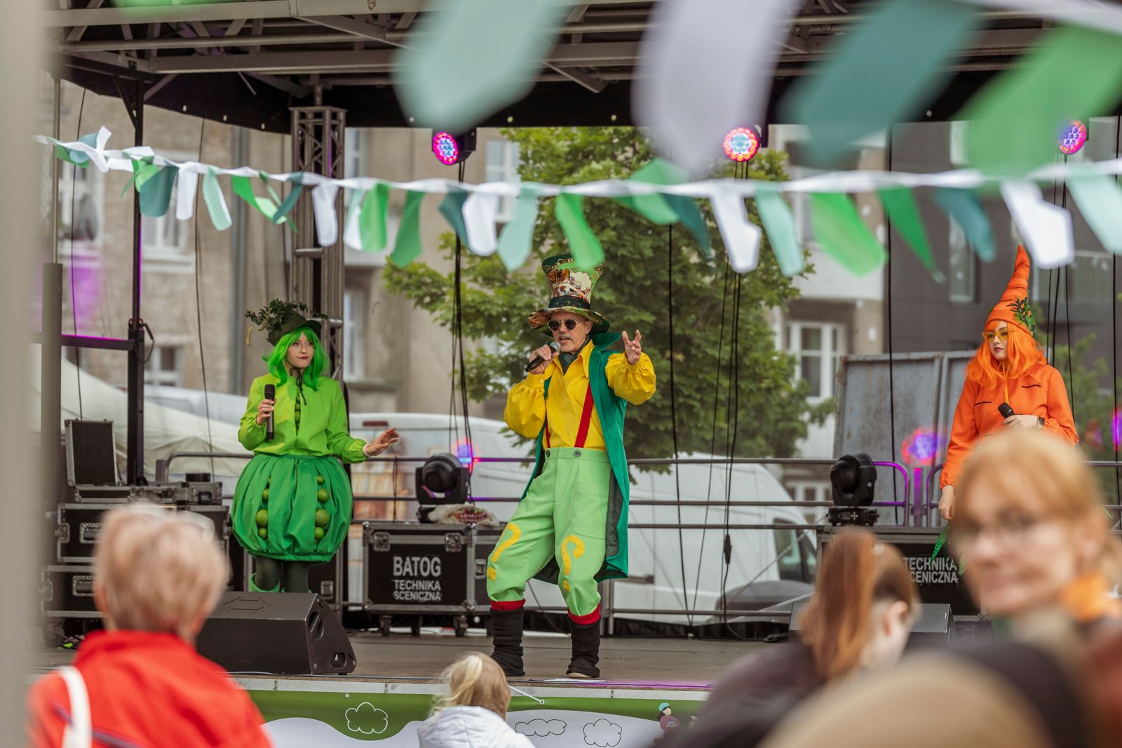 Zdjęcie przedstawia przebranych artystów - dwie kobiety i mężczyznę na scenie. Kobieta śpiewa do mikrofonu.
