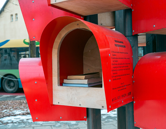 Zdjęcie przedstawia regał z książkami na zewnątrz. Ma on ciekawy kształt, połączonych małych budek.