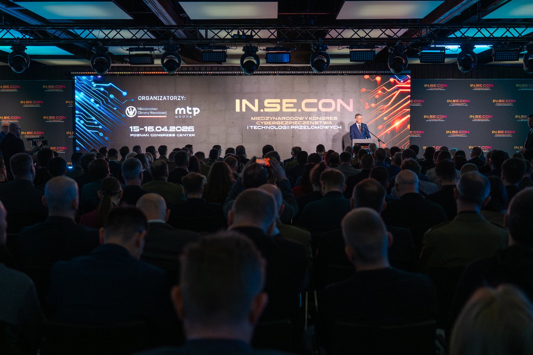 Zdjęcie przedstawia salę konferencyjną podczas otwarcia Międzynarodowego Kongresu Cyberbezpieczeństwa i Technologii Przełomowych IN.SE.CON w Poznań Congress Center. Na scenie, przed dużym ekranem z logo wydarzenia i datą 15-16.04.2026, przemawia mężczyzna w garniturze. Widownia jest wypełniona uczestnikami, wśród których widać osoby w mundurach oraz cywilach, siedzące tyłem do obiektywu. Nad sceną zamontowane jest profesjonalne oświetlenie eventowe.