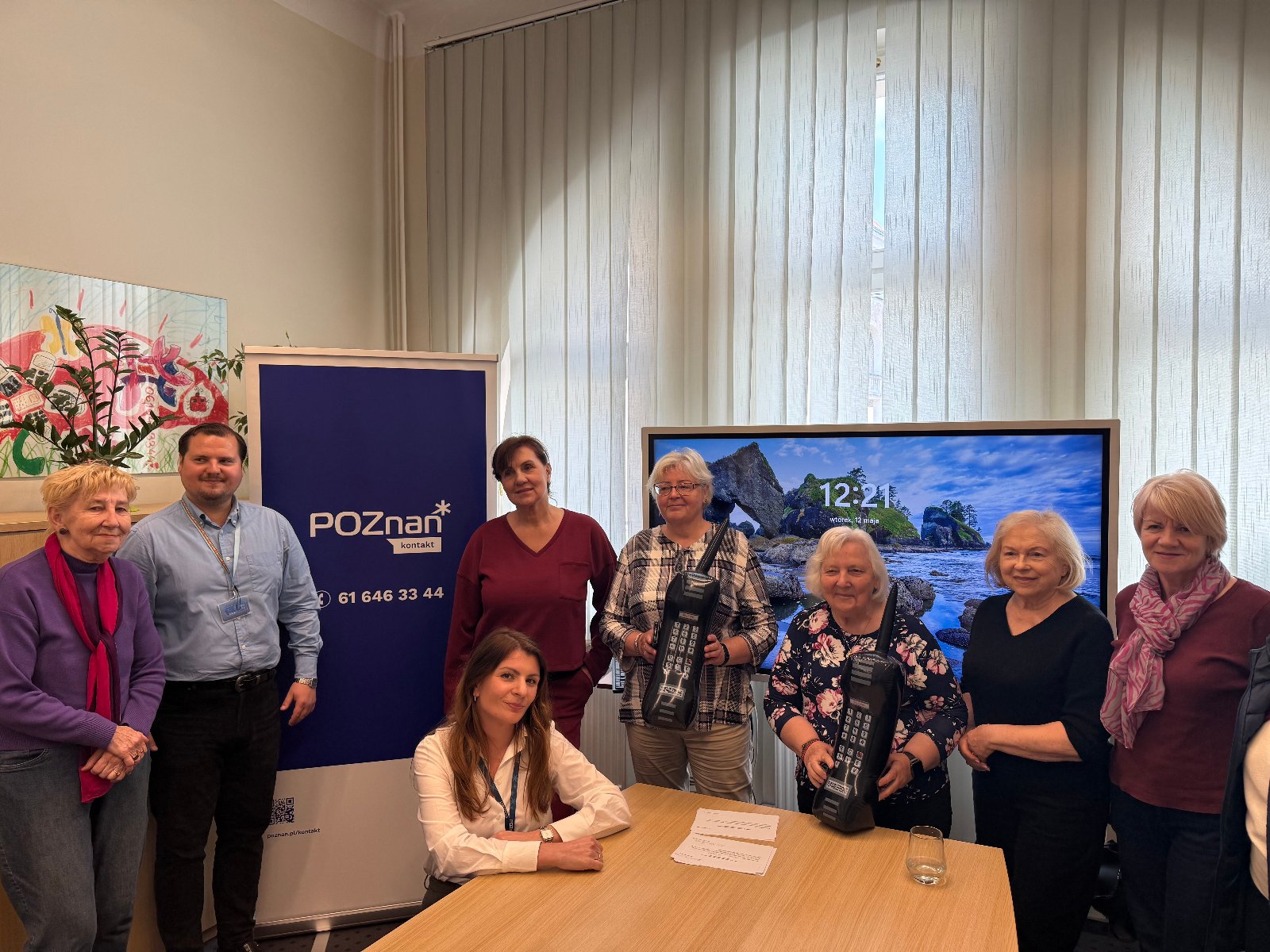 Zdjęcie przedstawia seniorki z poznańskich klubów seniora i wolontariuszki z Centrum Inicjatyw Senioralnych oraz pracowników Biura Poznań Kontakt na współnym pamiątkowym zdjęciu w siedzibie Biura, na zdjęciu widoczny jest również niebieski rollup z numerem telefony 61 646 33 44.
