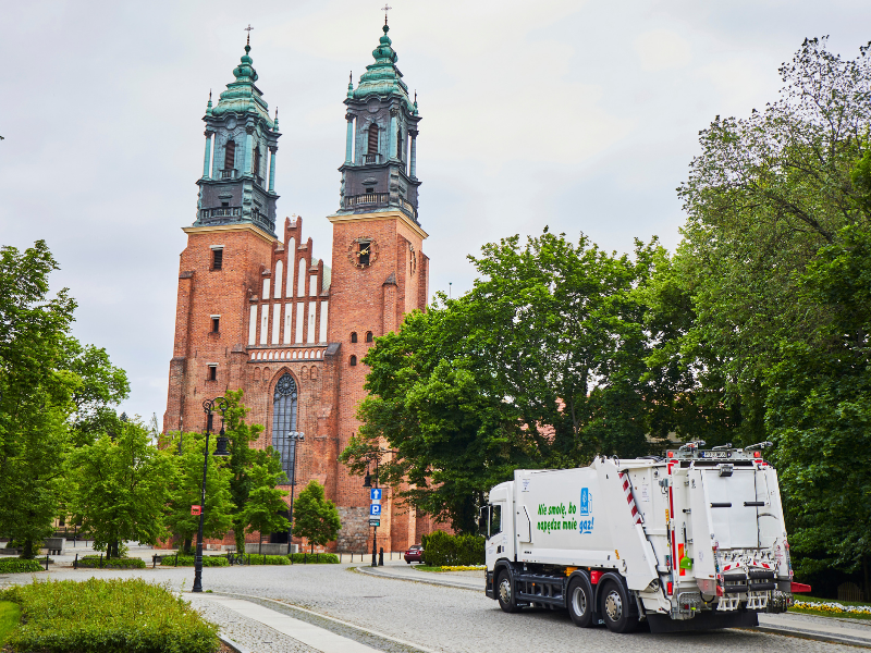 Zdjęcie przedstawia śmieciarkę jadącą po ulicy. Widać też katedrę i zieleń.