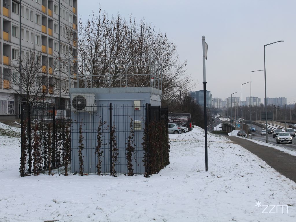 Zdjęcie przedstawia stację pomiaru jakości powietrza. Wokół niej leży śnieg. Widać też blok oraz tablicę z nazwą skweru.
