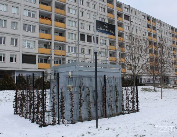 Zdjęcie przedstawia stację pomiaru jakości powietrza. Wokół niej leży śnieg. Widać też blok oraz tablicę z nazwą skweru.