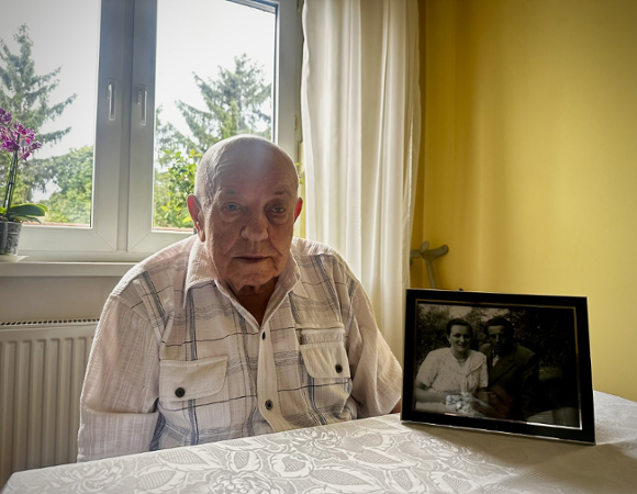 Zdjęcie przedstawia starszego mężczyznę siedzącego przy stole. Na stole obok niego stoi czarno białe zdjęcie mężczyzny i kobiety.