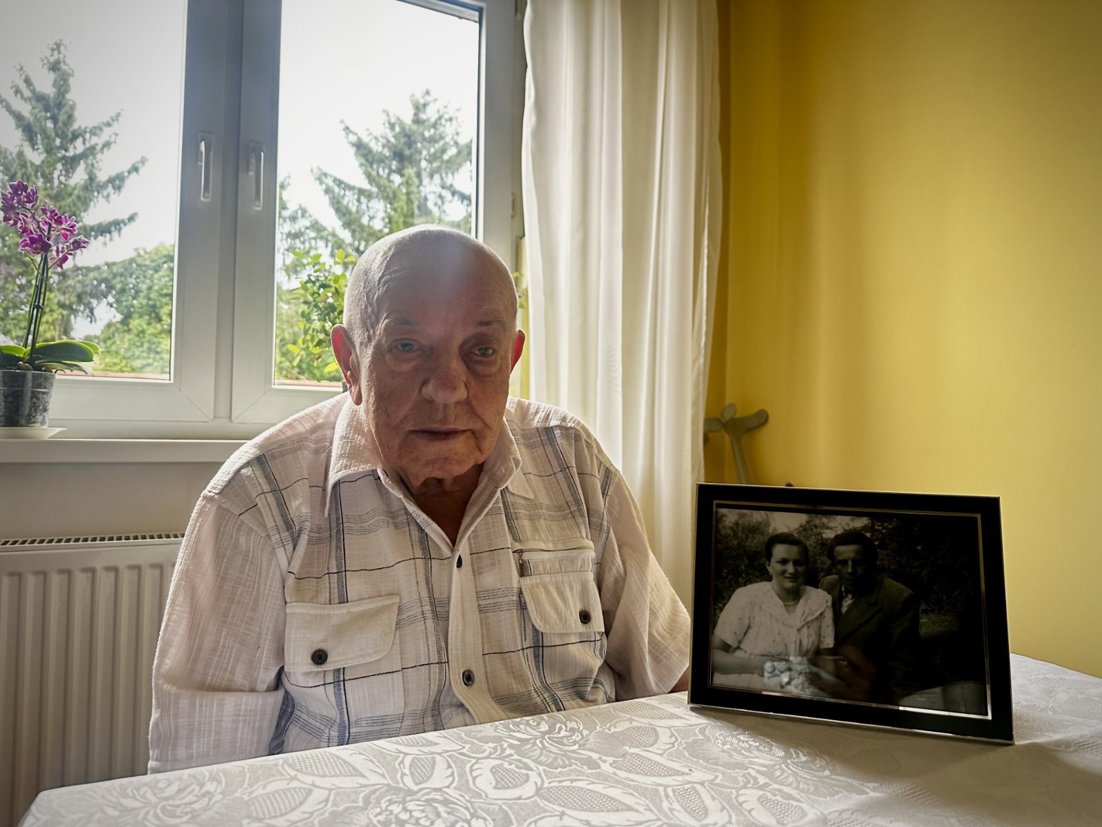 Zdjęcie przedstawia starszego mężczyznę siedzącego przy stole. Na stole obok niego stoi czarno białe zdjęcie mężczyzny i kobiety.