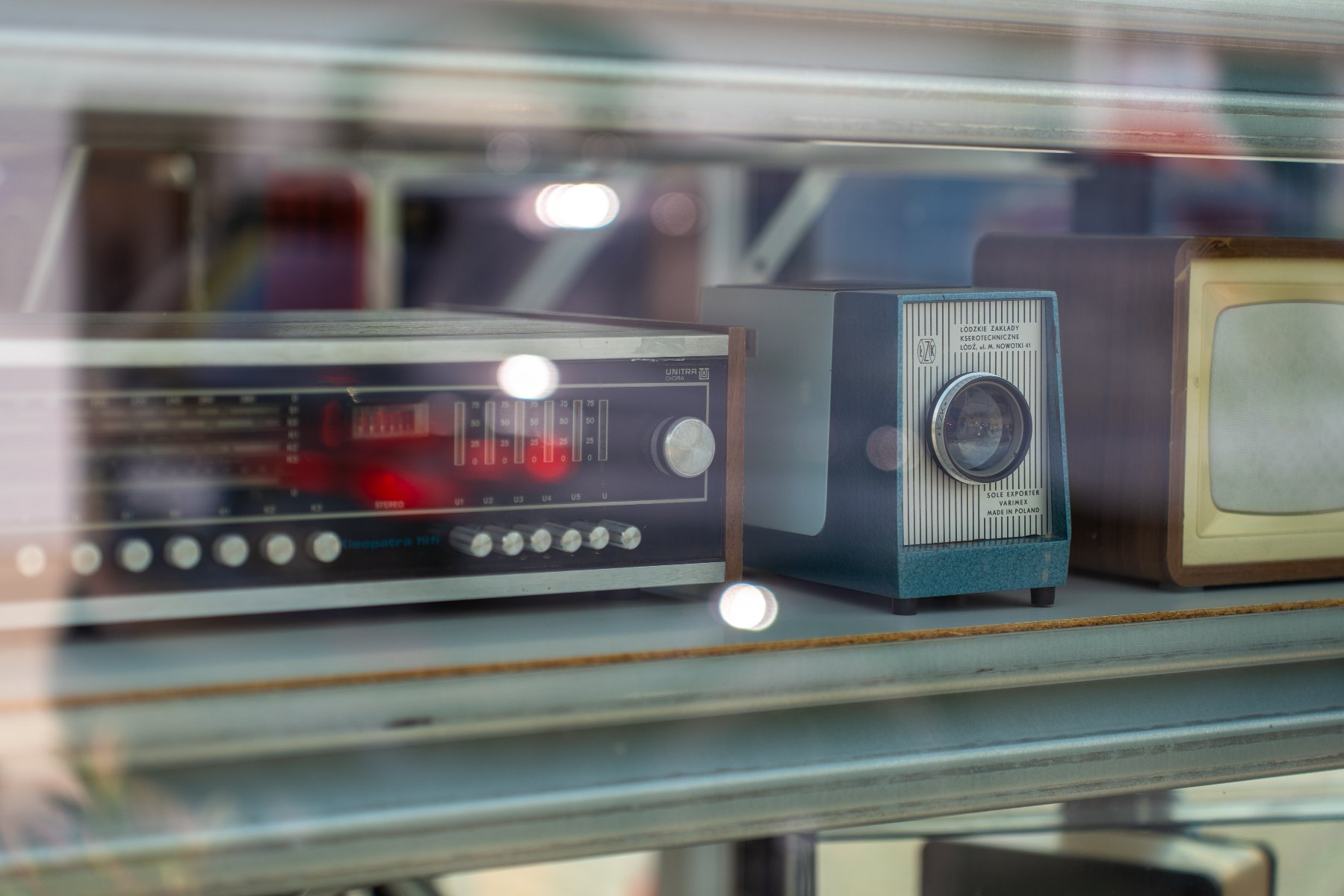Zdjęcie przedstawia stary sprzęt elektroniczny, radio, aparat, mały telewizorek, na półce za szybą