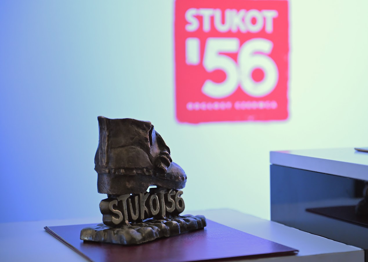 Zdjęcie przedstawia statuetkę nagrody Stukot '56 umieszczoną na ciemnoczerwonej podkładce na białym blacie. Statuetka ma formę odlanego z ciemnego metalu buta roboczego (tzw. "okuloka") o wyraźnej fakturze, osadzonego na podstawie z wypukłym napisem "STUKOT 56". W tle, na jasnej ścianie, widoczne jest rozmyte, czerwone logo z białym napisem "STUKOT '56".