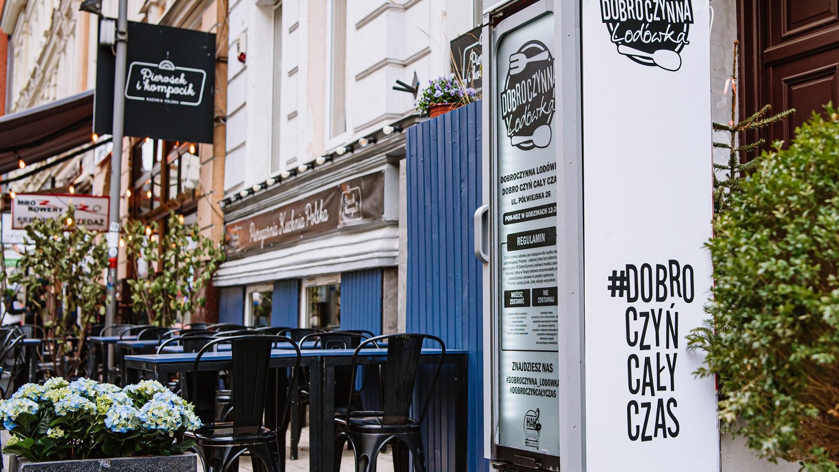 Zdjęcie przedstawia stojącą na ulicy lodówkę dobroczynną. Obok widać stoliki i krzesła.