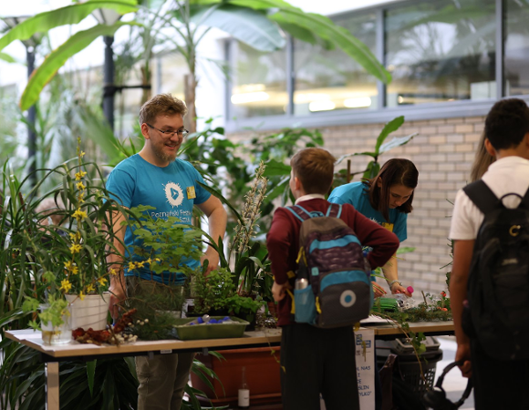 Zdjęcie przedstawia stół, na którym stoją rośliny oraz rozmawiających naprzeciwko siebie mężczyznę i chłopca.
