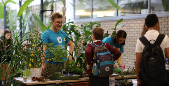 Zdjęcie przedstawia stół, na którym stoją rośliny oraz rozmawiających naprzeciwko siebie mężczyznę i chłopca.