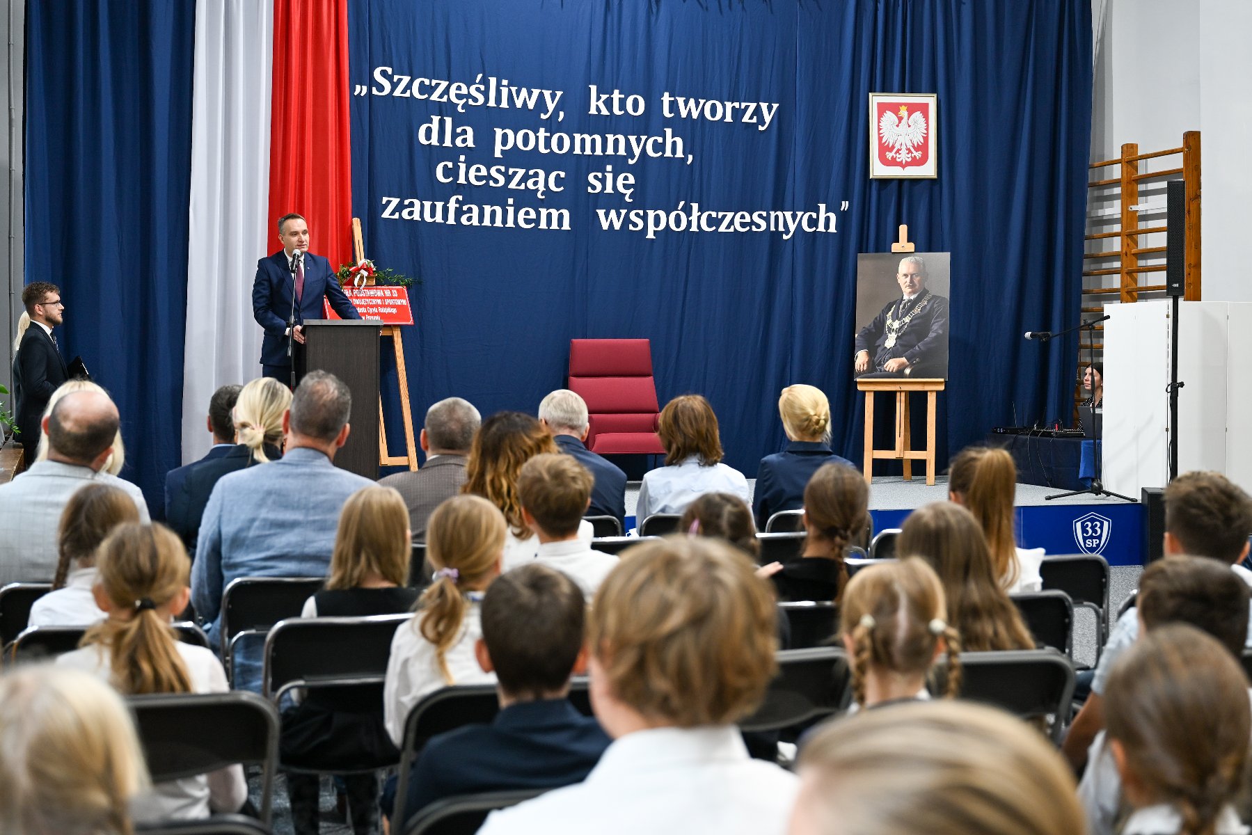 Zdjęcie przedstawia tabliczkę z nazwą szkoły i jej patronem stojącą na sztalugach i udekorowaną zielonymi gałązkami i wstążką.