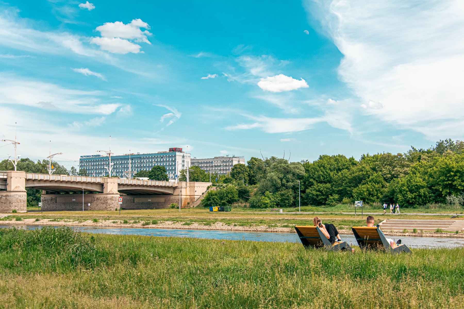Zdjęcie przedstawia tereny nad rzeką, ludzi na leżakach, w oddali też most i budynki.