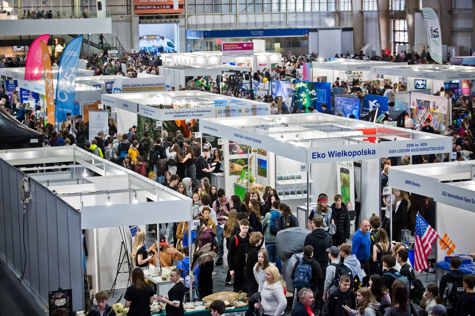 Zdjęcie przedstawia trójkę młodych ludzi i robota. Obok widać innych młodych ludzi.