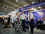 Zdjęcie przedstawia trójkę młodych ludzi i robota. Obok widać innych młodych ludzi.