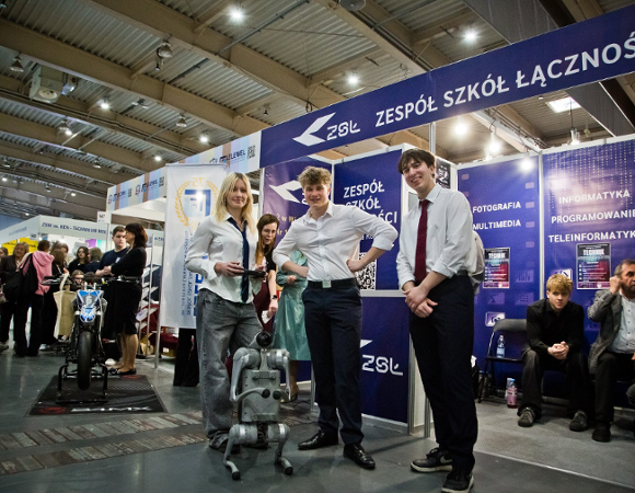 Zdjęcie przedstawia trójkę młodych ludzi i robota. Obok widać innych młodych ludzi.