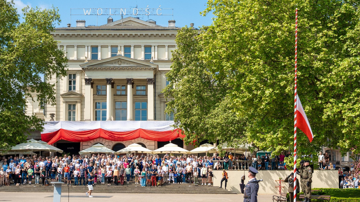 Zdjęcie przedstawia uroczyste obchody państwowe na Placu Wolności w Poznaniu. Na tle zabytkowego budynku, ozdobionego biało-czerwoną flagą oraz napisem "wolność" na dachu, zgromadził się tłum mieszkańców. Po prawej stronie widoczny jest wysoki maszt w biało-czerwone pasy, na który wciągana jest polska flaga przez żołnierzy w umundurowaniu galowym. Całość odbywa się w słoneczny dzień w otoczeniu zielonych drzew.
