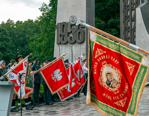 Zdjęcie przedstawia uroczystości patriotyczne odbywające się przed Pomnikiem Poznańskiego Czerwca 1956. Na pierwszym planie widoczny jest poczet sztandarowy z bogato zdobionym czerwonym sztandarem z wizerunkiem Matki Boskiej. W tle stoją rzędem harcerze oraz przedstawiciele służb mundurowych trzymający polskie flagi narodowe. Nad zgromadzonymi góruje fragment betonowego pylonu pomnika z wyraźną datą "1956".