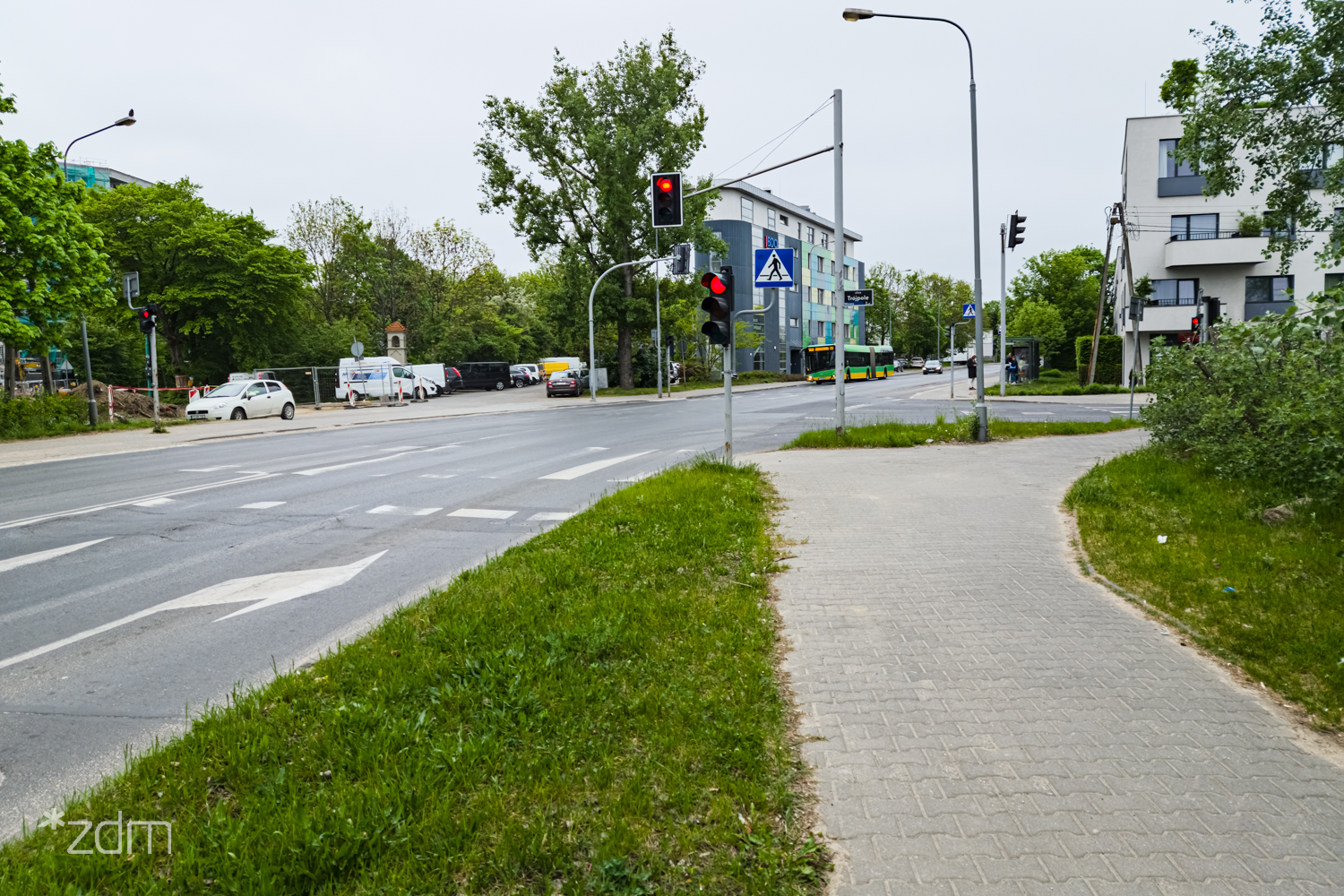 Zdjęcie przedstawia widok na miejskie skrzyżowanie z perspektywy chodnika wykonanego z kostki brukowej. Na pierwszym planie widać pas zieleni oddzielający drogę od ścieżki dla pieszych, a na jezdni namalowane są strzałki kierunkowe.