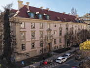 Zdjęcie przedstawia widok z góry na monumentalną kamienicę o jasnoróżowej elewacji i spadzistym, ceglanym dachu z lukarnami. Budynek posiada bogato zdobione obramowania okienne oraz dekoracyjny ryzalit z balkonem nad wejściem głównym. Przed kamienicą przebiega ulica z zaparkowanymi samochodami, a wzdłuż niej rosną drzewa, w tym jedno o wyraźnie żółtych, wiosennych liściach.