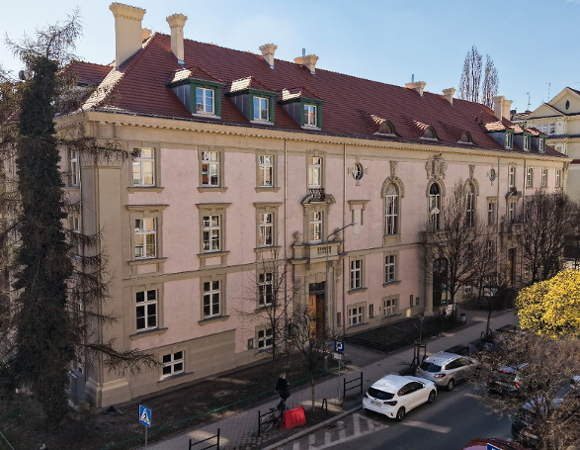 Zdjęcie przedstawia widok z góry na monumentalną kamienicę o jasnoróżowej elewacji i spadzistym, ceglanym dachu z lukarnami. Budynek posiada bogato zdobione obramowania okienne oraz dekoracyjny ryzalit z balkonem nad wejściem głównym. Przed kamienicą przebiega ulica z zaparkowanymi samochodami, a wzdłuż niej rosną drzewa, w tym jedno o wyraźnie żółtych, wiosennych liściach.