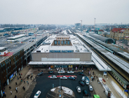 Zdjęcie przedstawia widok z lotu ptaka na stary budynek dworca kolejowego Poznań Główny w zimowej scenerii. Na dachu obiektu, przykrytym cienką warstwą śniegu, widnieje duży niebieski napis z nazwą stacji, a przed wejściem znajduje się półkolisty podjazd, na którym parkują liczne taksówki.