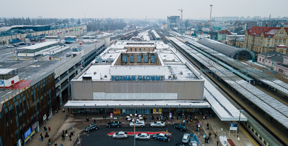 Zdjęcie przedstawia widok z lotu ptaka na stary budynek dworca kolejowego Poznań Główny w zimowej scenerii. Na dachu obiektu, przykrytym cienką warstwą śniegu, widnieje duży niebieski napis z nazwą stacji, a przed wejściem znajduje się półkolisty podjazd, na którym parkują liczne taksówki.