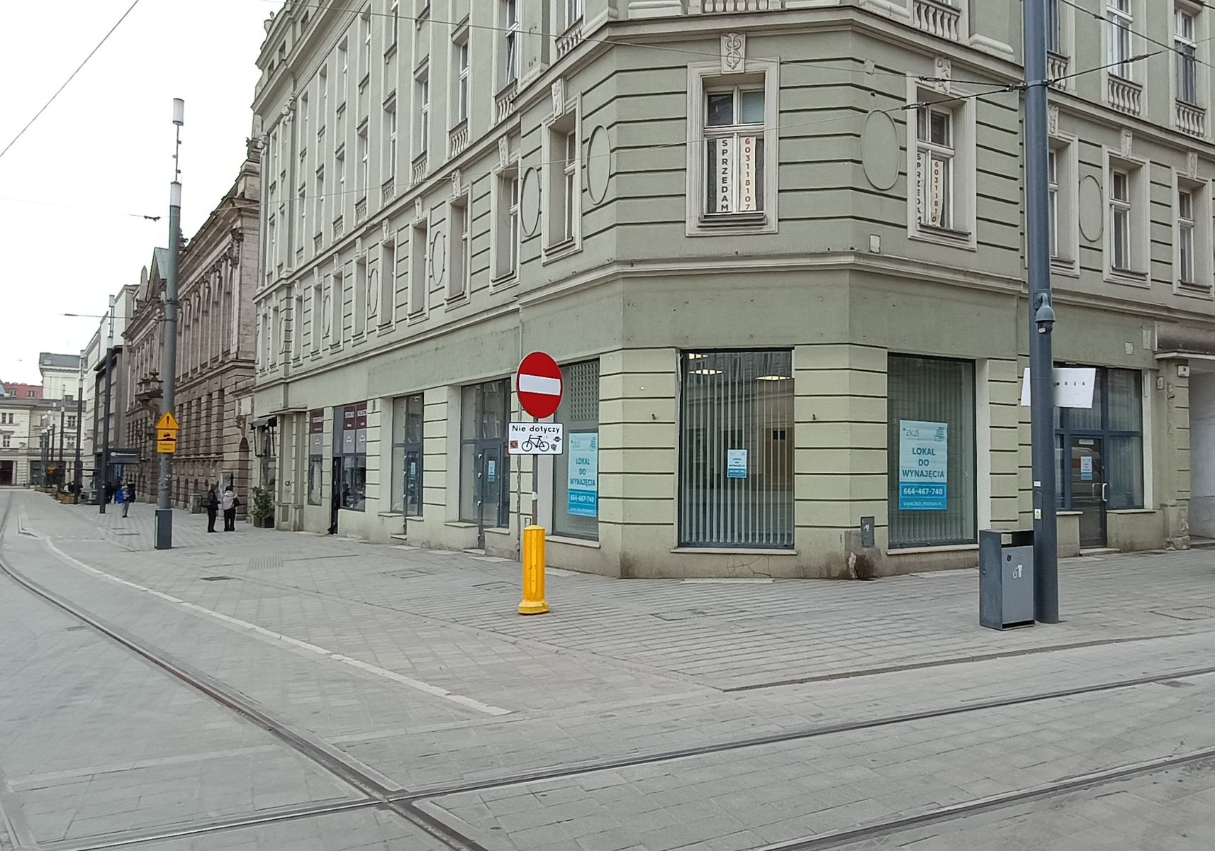 Zdjęcie przedstawia widok z poziomu ulicy na narożnik zabytkowej, kilkupiętrowej kamienicy o jasnoszarej elewacji. W parterze budynku znajdują się duże witryny sklepowe, na których naklejono niebiesko-białe plakaty informacyjne z napisem ZKZK "LOKAL DO WYNAJĘCIA" oraz numerem telefonu.