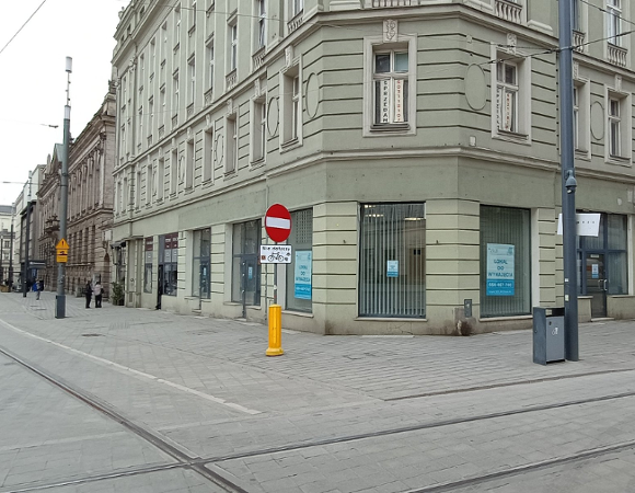 Zdjęcie przedstawia widok z poziomu ulicy na narożnik zabytkowej, kilkupiętrowej kamienicy o jasnoszarej elewacji. W parterze budynku znajdują się duże witryny sklepowe, na których naklejono niebiesko-białe plakaty informacyjne z napisem ZKZK "LOKAL DO WYNAJĘCIA" oraz numerem telefonu.