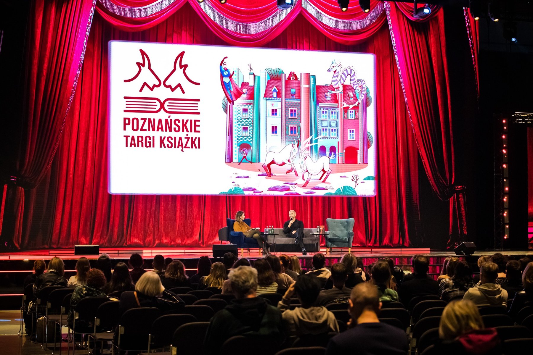 Zdjęcie przedstawia wielki stół, na którym znajdują się budowle z klocków oraz oglądających je ludzi.