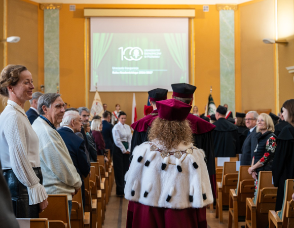 Zdjęcie przedstawia władze uczelni idące korytarzem pomiędzy rzędami krzeseł.