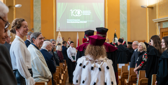 Zdjęcie przedstawia władze uczelni idące korytarzem pomiędzy rzędami krzeseł.