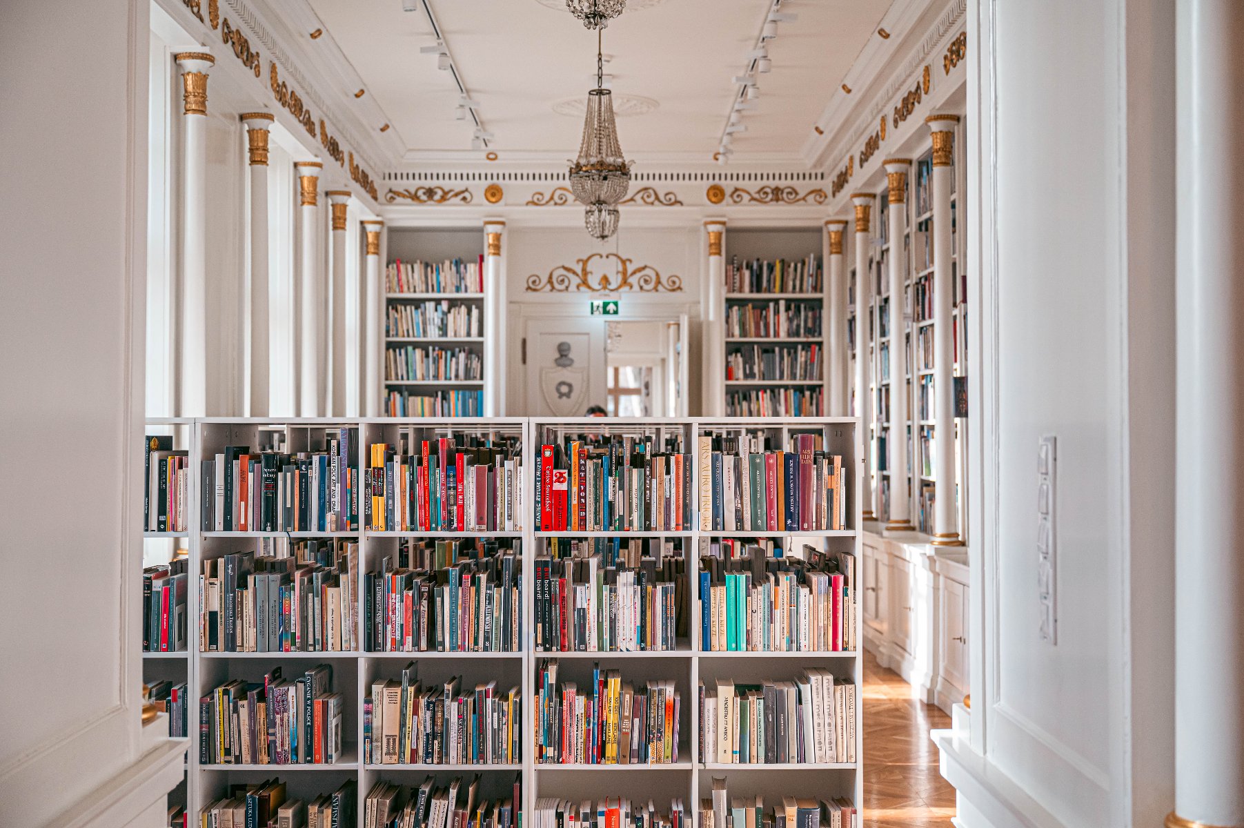 Zdjęcie przedstawia wnętrze eleganckiej, klasycystycznej biblioteki. W centrum kadru znajduje się biały regał wypełniony różnokolorowymi książkami, który dzieli przestrzeń. W tle widać dalszą część pomieszczenia z wysokimi regałami sięgającymi sufitu, ozdobionymi złotymi detalami i kolumnami z korynckimi głowicami. Nad przejściem widnieje dekoracyjny ornament, a z sufitu zwisa kryształowy żyrandol.