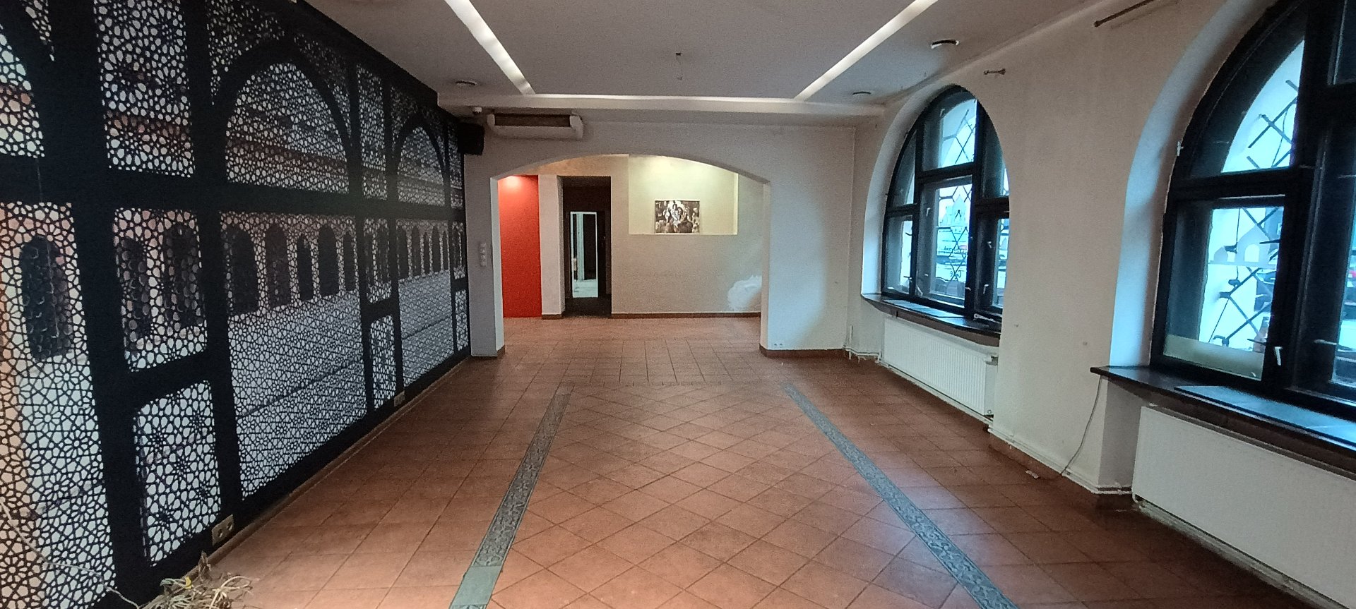 Zdjęcie przedstawia wnętrze pustego lokalu użytkowego, prawdopodobnie przeznaczonego pod gastronomię lub galerię. Po lewej stronie dominuje ściana z czarną fototapetą przedstawiającą ażurowe, orientalne łuki. Podłoga wyłożona jest terakotą w ciepłym odcieniu, z ozdobnym ciemniejszym pasem biegnącym wzdłuż sali. Po prawej stronie znajdują się dwa duże, łukowate okna z widokiem na ulicę.