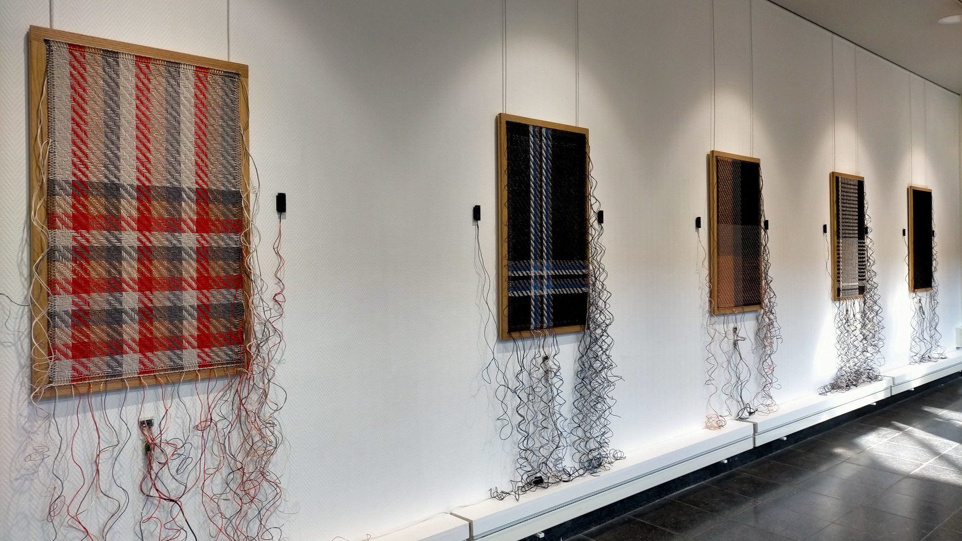 Zdjęcie przedstawia wystawę autorstwa Cécile Babiole w jasnej galerii. Na białej ścianie zawieszono w rzędzie pięć prostokątnych prac w drewnianych ramach, które przypominają tkaniny o różnych wzorach (między innymi we wzór kratki w kolorach czerwonym, niebieskim i czarnym). Z dołu każdej ramy swobodnie opadają na podłogę liczne, splątane kable i przewody, tworząc efekt "rozpruwania się" dzieł.