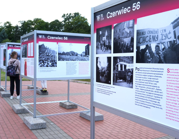 Zdjęcie przedstawia wystawę z opisem i zdjęciami Poznańskiego Czerwca. Wokół widać zieleń, staw i spacerujących ludzi.