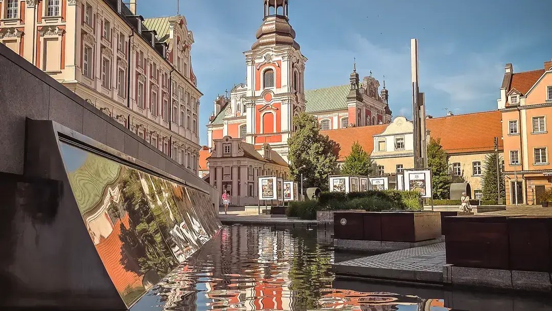 Zdjęcie przedstawia zabytkowy plac miejski otoczony pastelowymi kamienicami. W centrum kadru dominuje kościół z wysoką wieżą. Odbicie kościoła widoczne jest w spokojnej tafli wody znajdującej się na pierwszym planie. Wokół placu znajdują się rabaty zieleni, plansze wystawowe i nowoczesne elementy małej architektury.