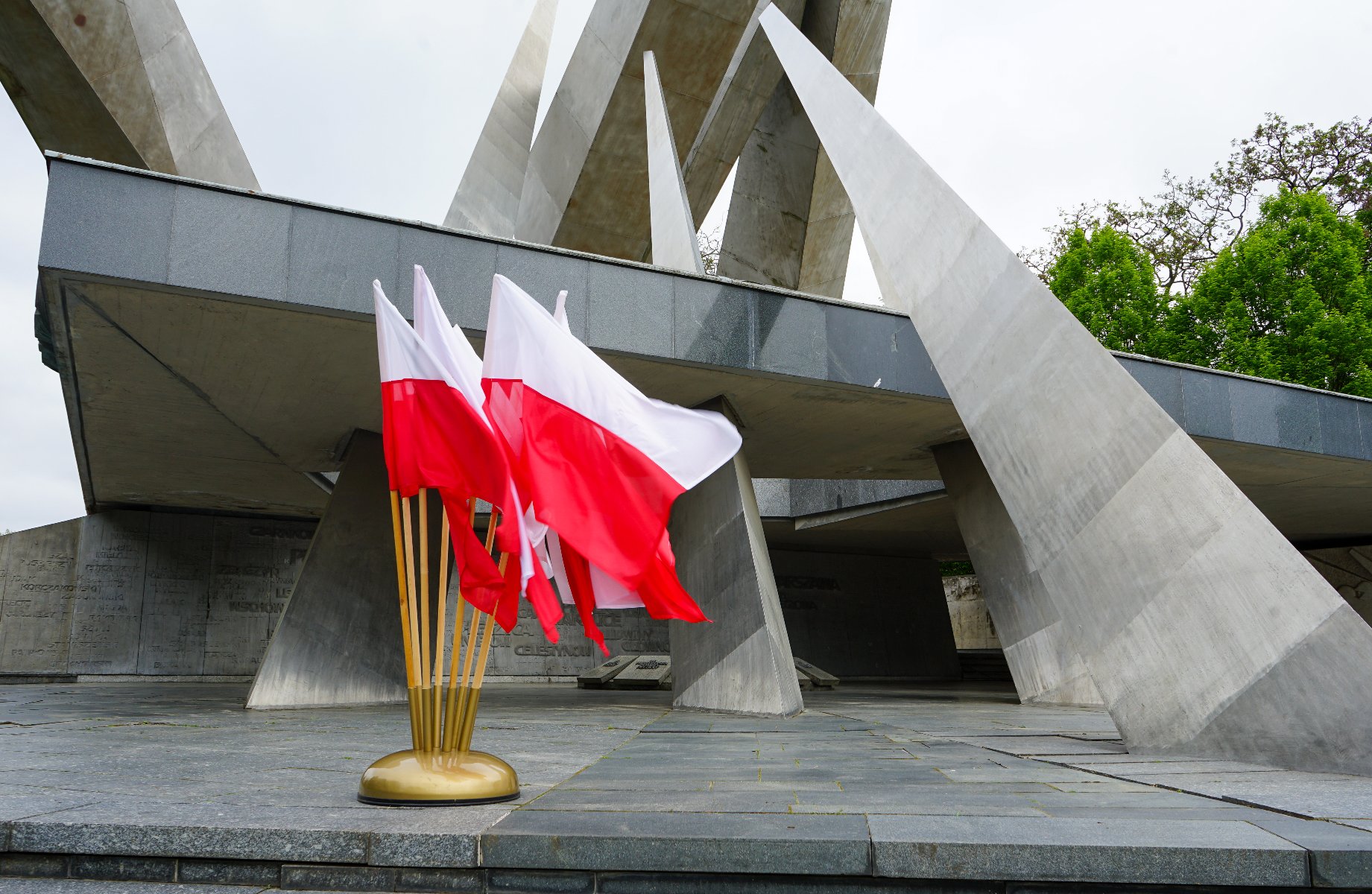 Zdjęcie przedstawia zbliżenie na bukiet białych i czerwonych flag narodowych Polski na złocistej podstawce, umieszczonych przed imponującym, nowoczesnym pomnikiem z betonu i metalu. W tle widać dynamiczną, geometryczną architekturę pomnika z ostrymi, wznoszącymi się elementami, kontrastującą z miękkimi fałdami flag.