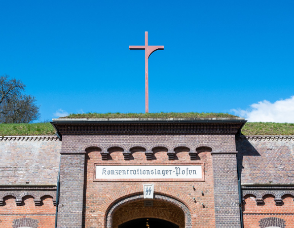 Zdjęcie przedstawia zbliżenie na górną część bramy wejściowej do historycznego obiektu z czerwonej cegły (Fort VII w Poznaniu). Nad łukiem bramy widnieje biała tablica z napisem "Konzentrationslager-Posen".