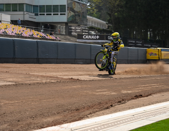 Zdjęcie przedstawia żużlowca w żółtym kombinezonie i kasku. Zawodnik jedzie na tylnym kole, wzbijając za sobą gęstą chmurę pyłu, co nadaje ujęciu dynamizmu. W tle widoczne są puste trybuny z żółto-różowymi krzesełkami oraz fragmenty infrastruktury sportowej i otaczająca stadion zieleń drzew.